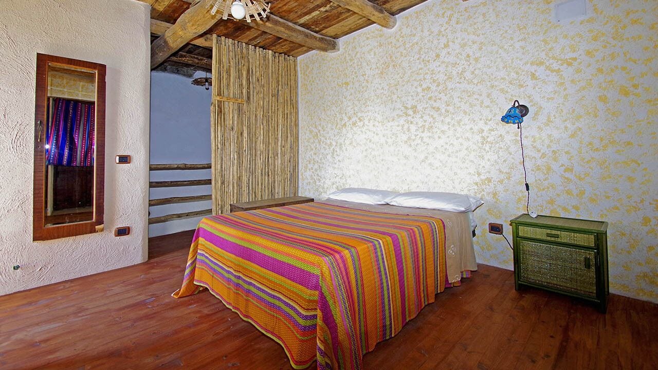A bedroom with a wooden floor, a bed with a colorful striped bedspread, a bedside table with a lamp, and a window with curtains.