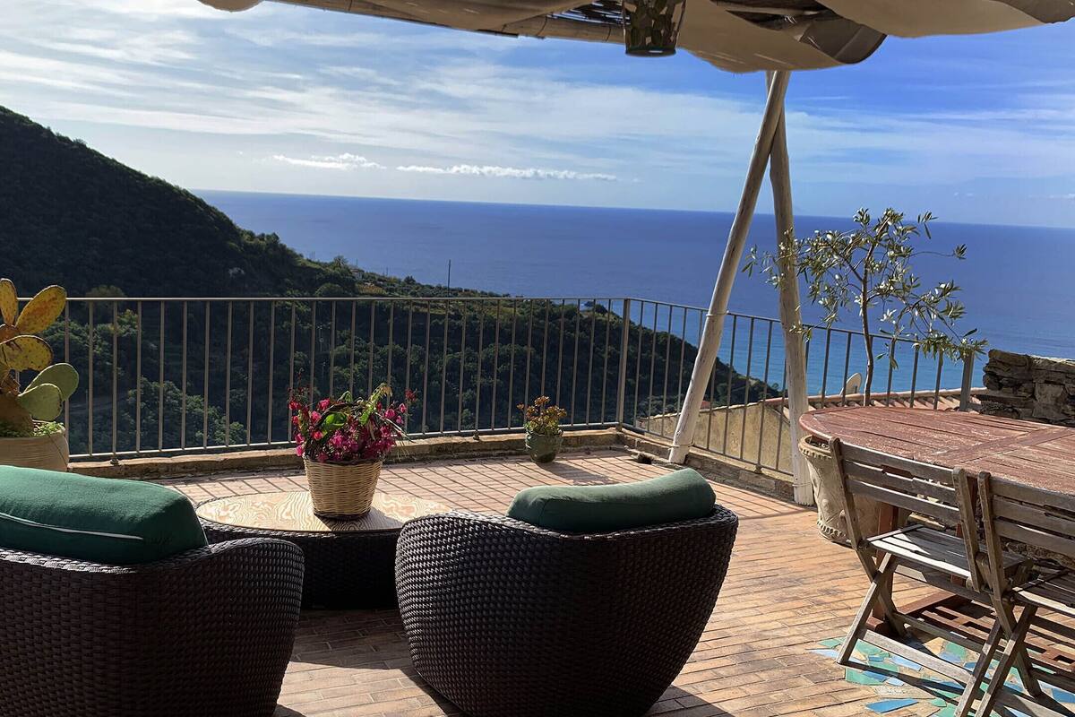 A terrace with wicker furniture, a table, and potted plants, overlooking a coastal landscape.