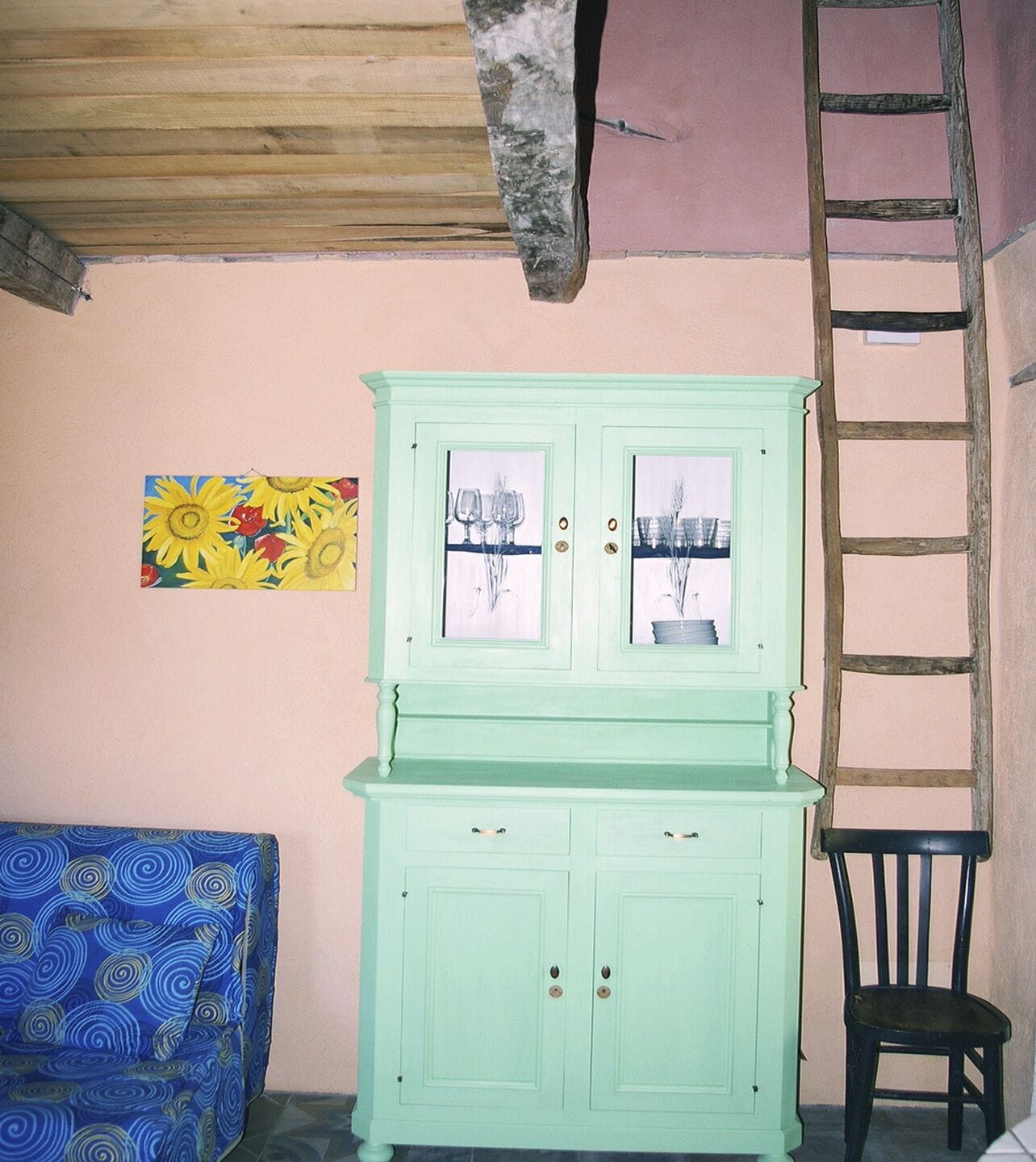 A room with a light green cabinet, a blue patterned sofa, a ladder, and a painting of sunflowers.