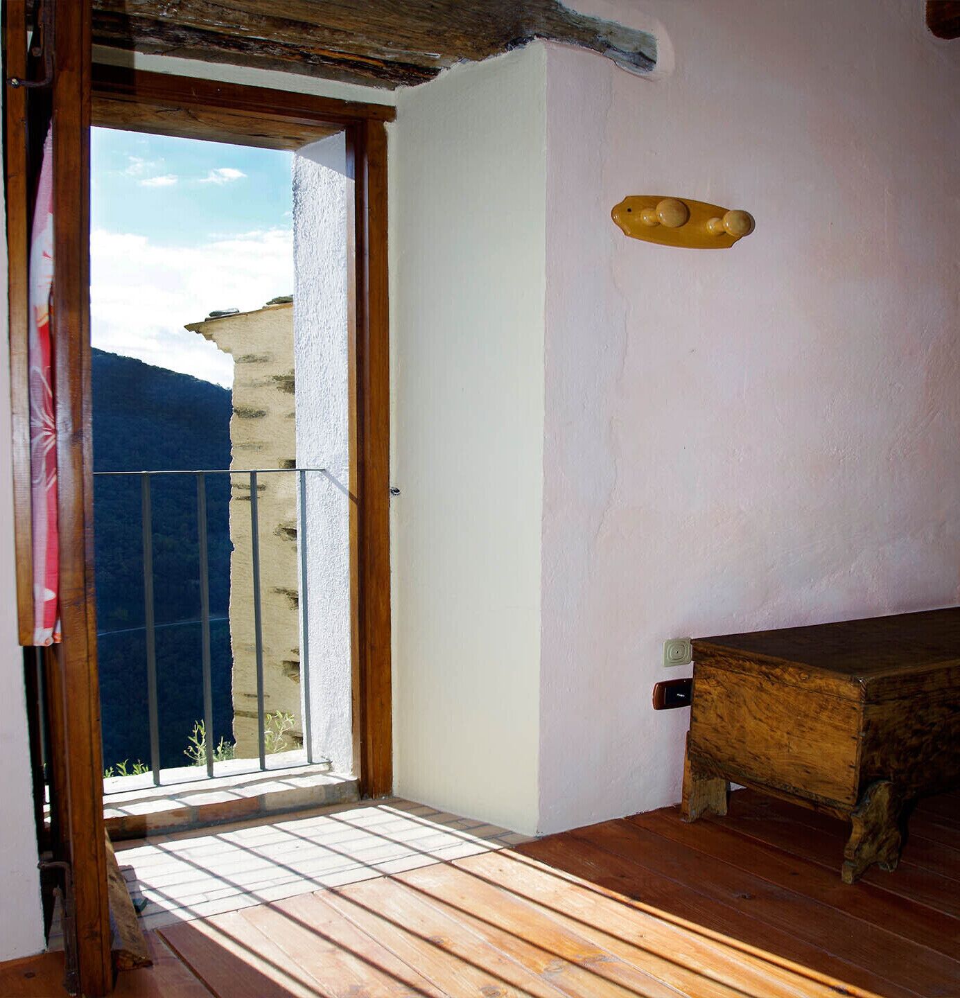 Een kamer met houten vloer, een houten bank, een witte muur en een balkon met uitzicht op bergen.