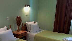 A hotel room with two beds, each with white pillows and green bedding. There is a nightstand with a vase of dried plants and a wall-mounted light. - Vicky's Guesthouse (Corfu)