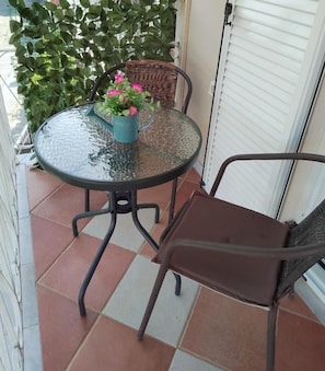 A round glass table with a metal base, accompanied by two wicker chairs, a potted plant with pink flowers, and a white door in the background. - Vicky's Guesthouse (Corfu)
