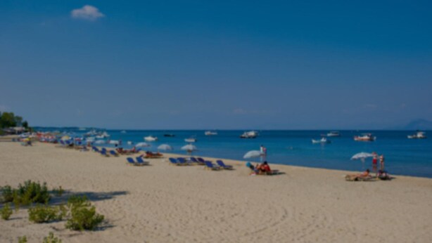 A beach with rows of sun loungers and umbrellas, a clear blue sky, and boats on the water. - Vicky's Guesthouse (Corfu)