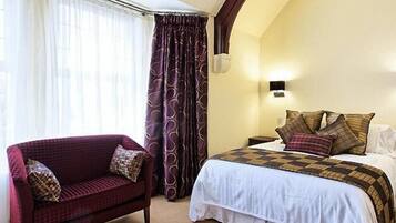 A hotel room with a bed, a red armchair, a nightstand, and a wall-mounted lamp.