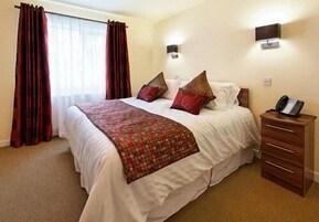 Quarto de hotel com uma cama grande, mesa de cabeceira de madeira, telefone e luminária fixada na parede.