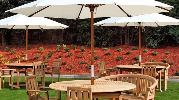 Houten tafels en stoelen buiten, met witte parasols, op een net gemaaid gazon met rood mulch landschap.