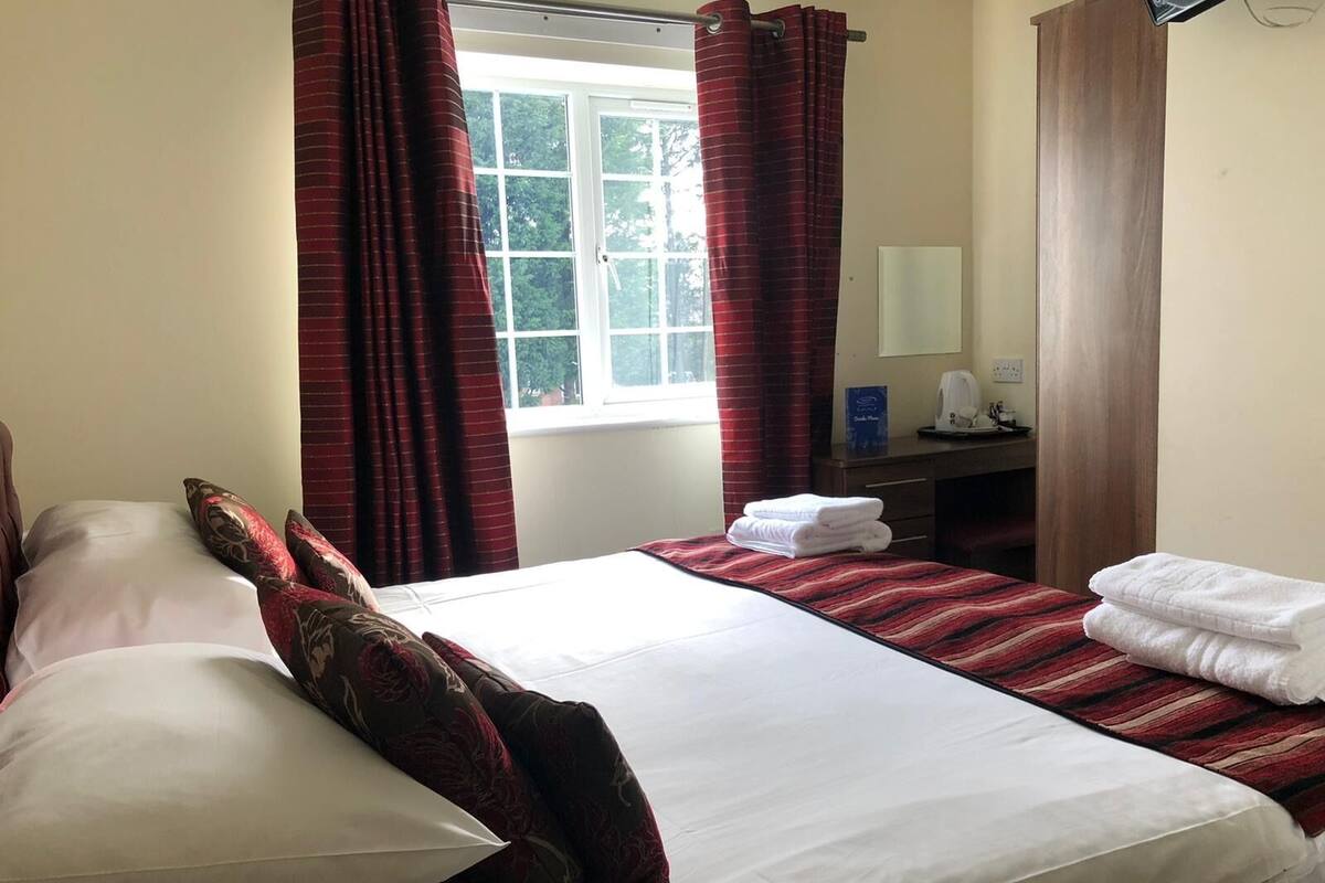 A hotel room with a large bed, two pillows, a bedside table with a lamp, and a window with curtains.