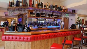 Een bar met meerdere flessen en glazen op de planken, een kassa en rode barstoelen.