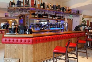 Uma área de bar com várias prateleiras de garrafas e copos, um caixa registradora e banquetas vermelhas.