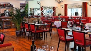 Een restaurant met rode bekleden zitbanken, houten tafels en een bar met wijnflessen.