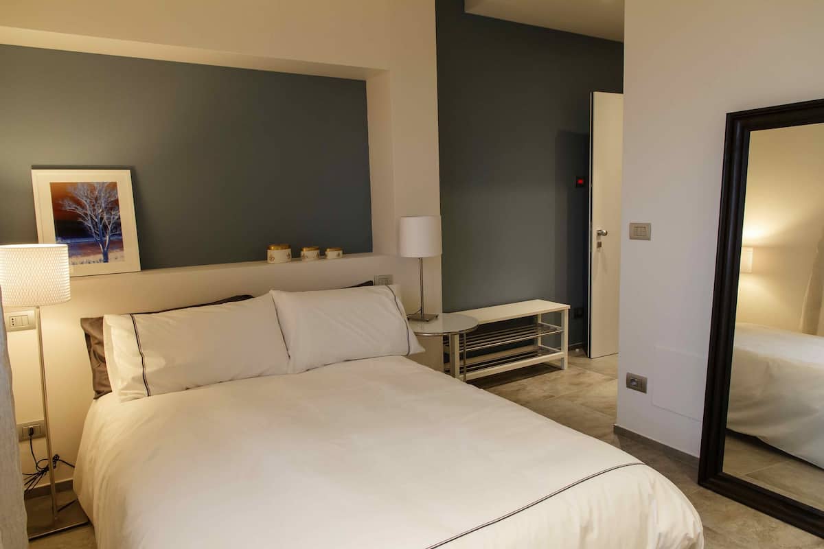A hotel room with a large bed, a bedside table, a lamp, a framed picture on the wall, and a mirror.