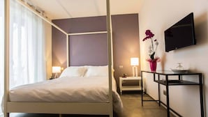 A modern hotel room with a four-poster bed, a flat-screen TV mounted on the wall, a bedside table with a lamp, and a small table with a vase of flowers. - B&B Cascina Belmonte (Fossano)
