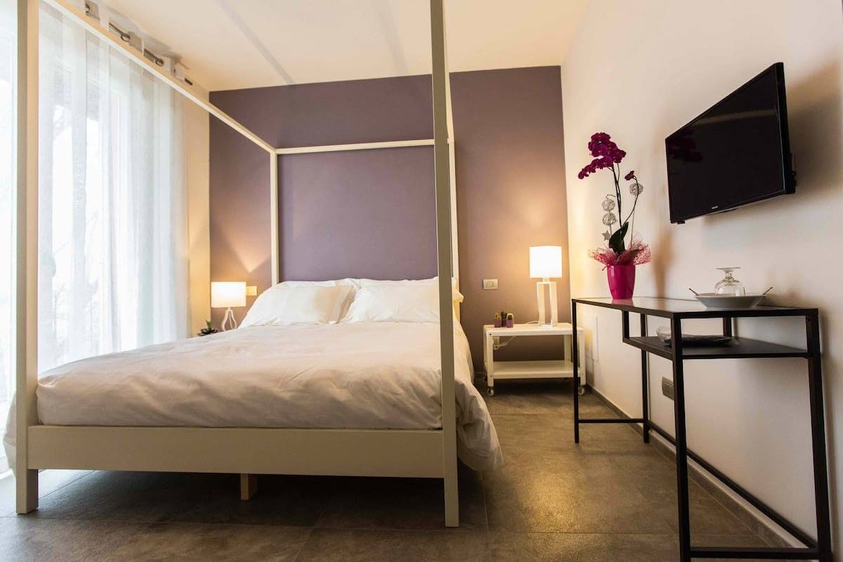 A modern hotel room with a four-poster bed, a flat-screen TV mounted on the wall, a bedside table with a lamp, and a small table with a vase of flowers.