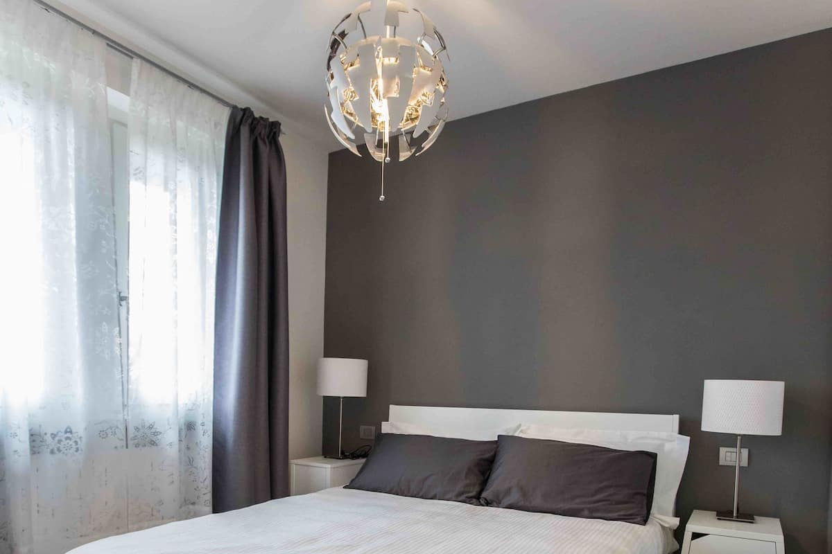 A modern bedroom with a large bed, two bedside tables with lamps, a chandelier, and sheer curtains.