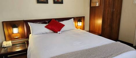 Una habitación de hotel con una cama grande, un armario de madera, una mesita de noche con teléfono e iluminación en la pared.