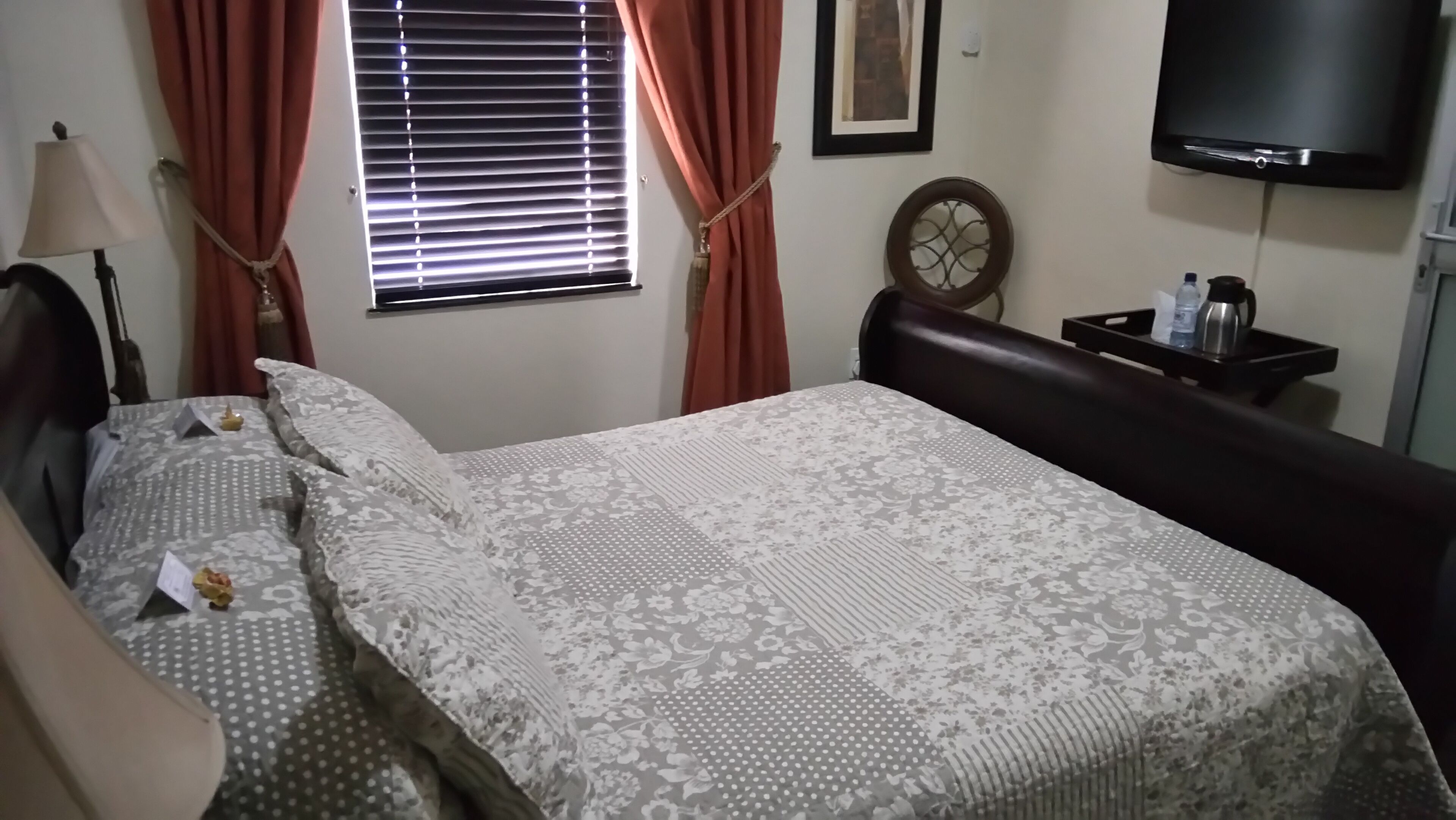 A neatly made bed with a patterned bedspread, a bedside lamp, a window with curtains, a wall-mounted TV, and a small table with a bottle and a kettle.
