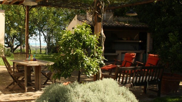 A patio area with a wooden table and chairs, surrounded by potted plants and a view of the sea. - Agriturismo La Barca nel Bosco (Fano)
