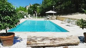 A rectangular swimming pool with a clear blue water surface, surrounded by a white tiled deck, and a few lounge chairs and umbrellas. - Agriturismo La Barca nel Bosco (Fano)