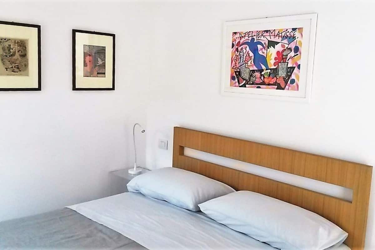 Une chambre à coucher avec une tête de lit en bois, deux œuvres d’art encadrées, une lampe de chevet et un lit recouvert de linge de maison blanc.