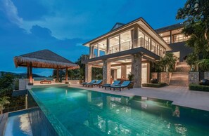 Una casa moderna de dos pisos con una gran piscina, un quiosco con techo de paja y varias sillas de descanso.
