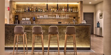 Una zona de bar con mostrador de madera, taburetes y una variedad de botellas y vasos.