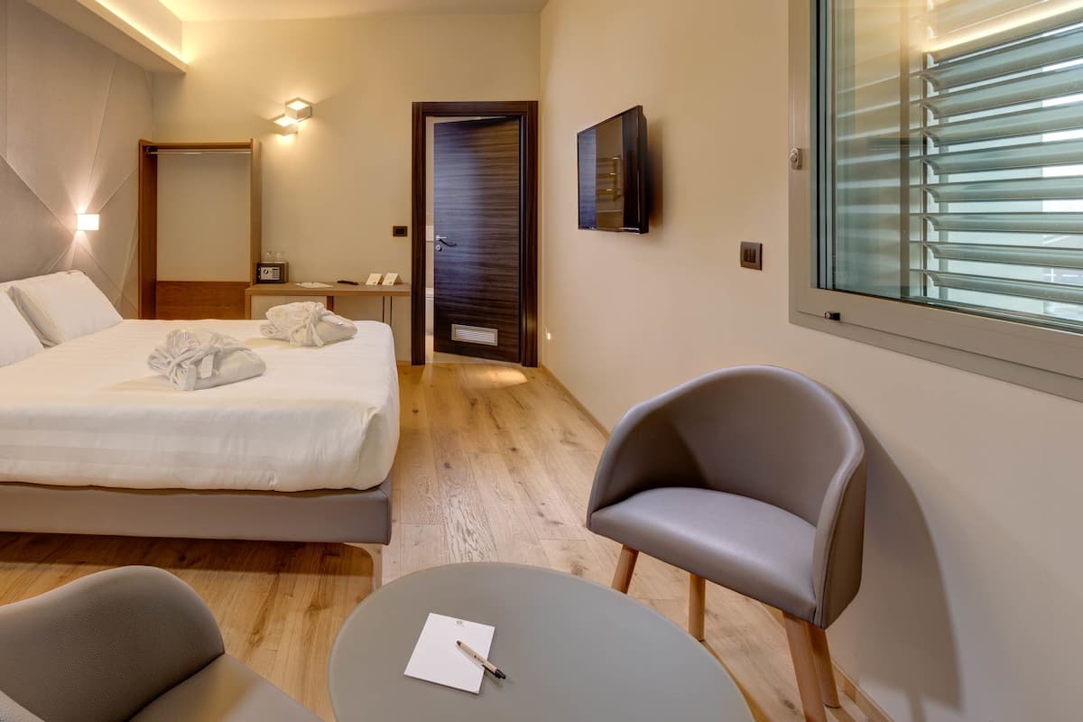 A modern hotel room with a neatly made bed, a sitting area with a chair and a small table, and a large window with blinds.