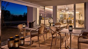 Un área de comedor al aire libre con sillas y mesas de mimbre, una zona de bar con mostrador y taburetes, y vista al espacio interior para comer.