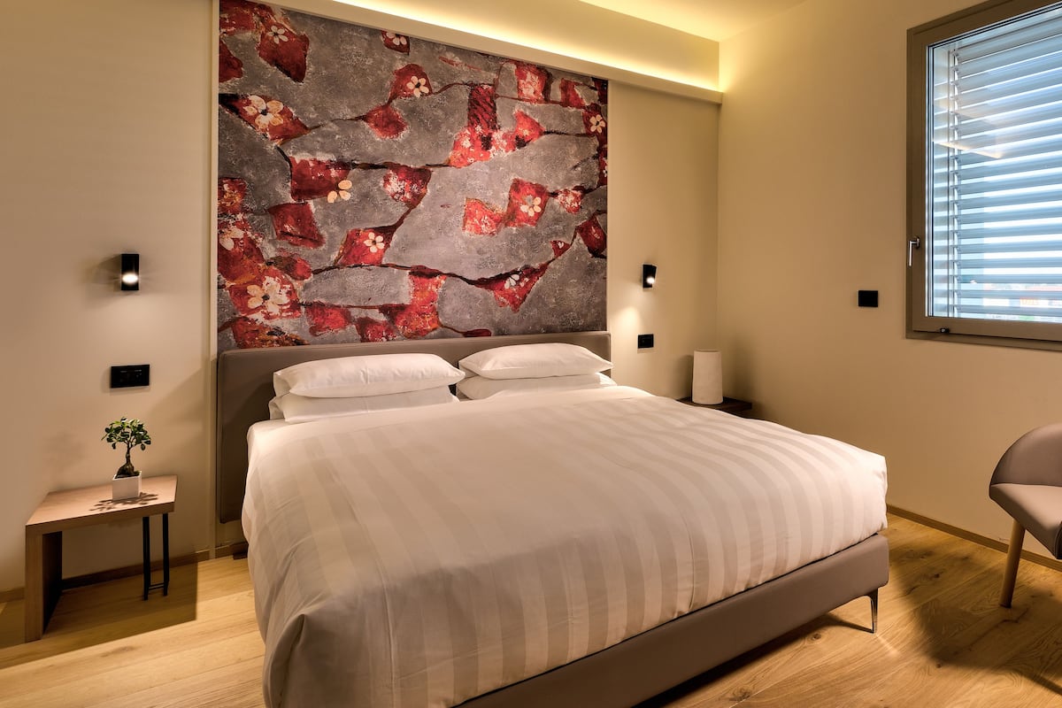 A modern bedroom with a large bed, a side table with a plant, a chair, and a large window with blinds.