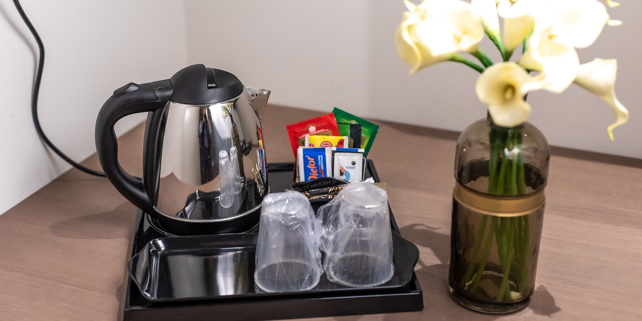A coffee maker, two glasses, a vase with flowers, and some packaged food on a tray.