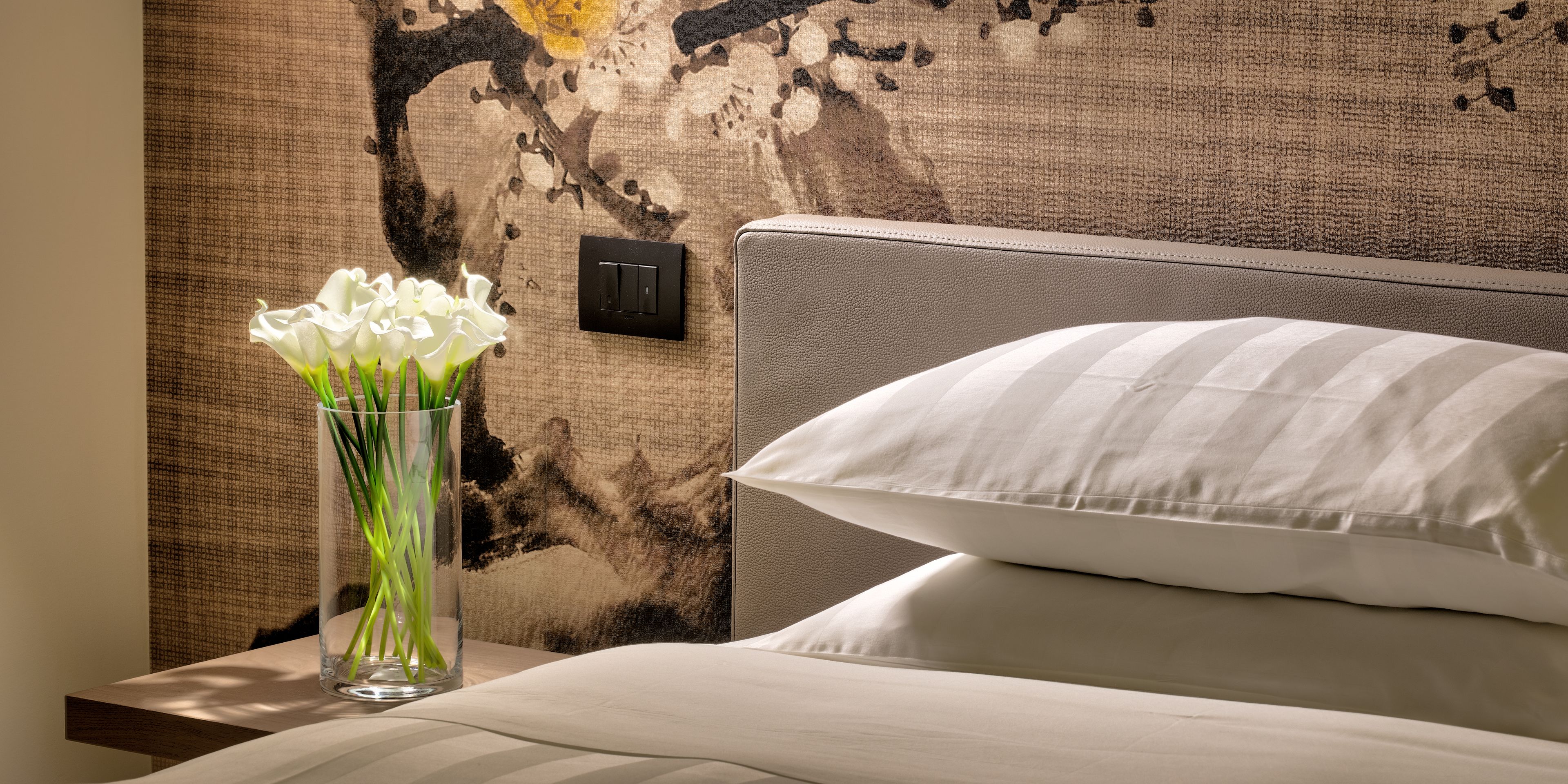 A hotel room with a bed, a nightstand, a vase with white flowers, and a textured wall with a switch.