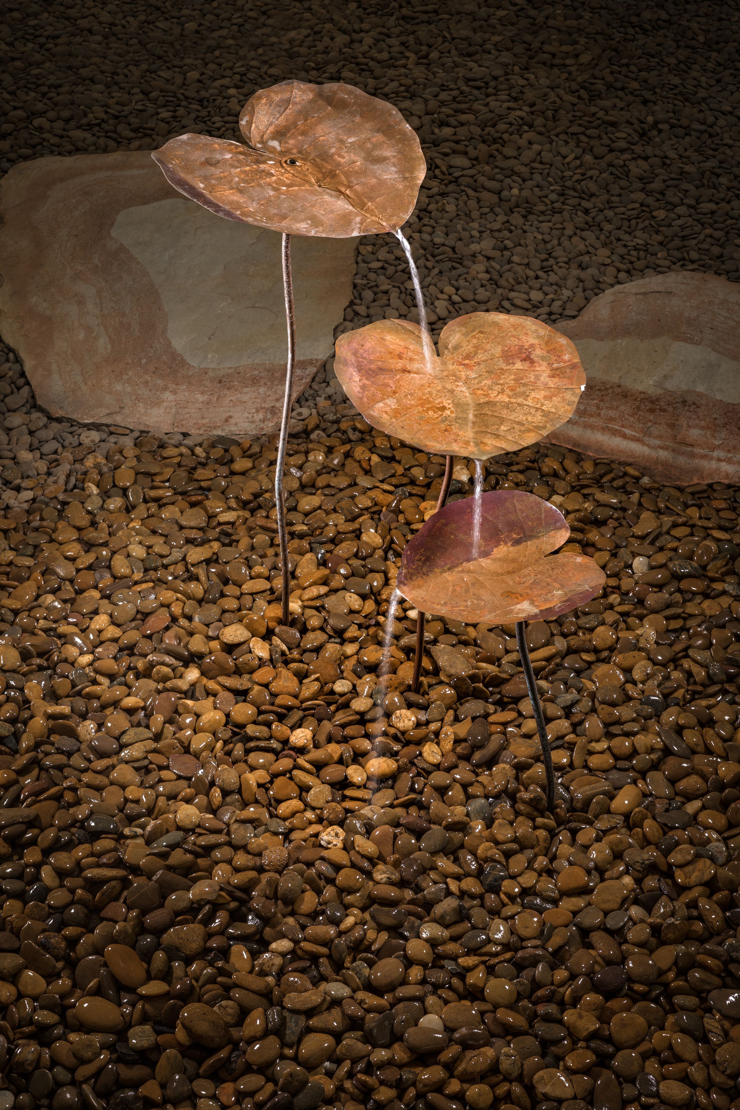 A water feature with two copper leaves and a stream of water flowing between them, set on a bed of pebbles.