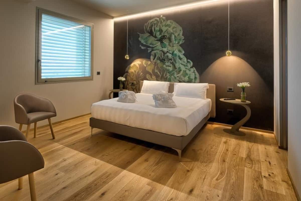 A modern bedroom with a wooden floor, a bed with white linens, a chair, a bedside table with a vase, and a large floral mural on the wall.