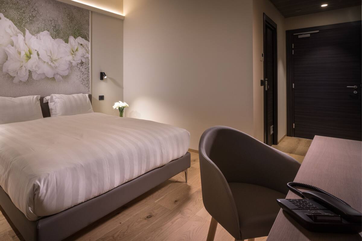 A modern bedroom with a large bed, bedside tables with lamps, a desk with a telephone, and a wall with a floral mural.