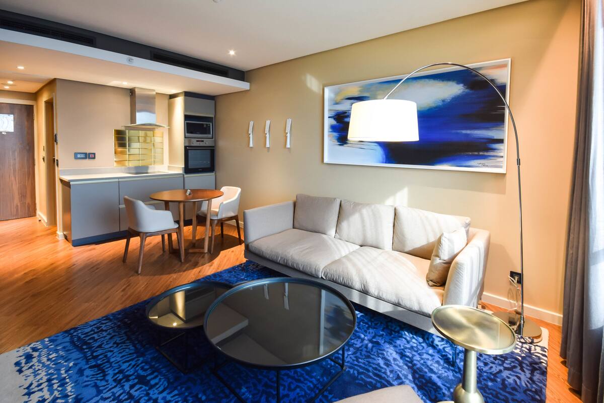 A modern living room with a sofa, two glass-top tables, a coffee table, and a dining area with chairs.