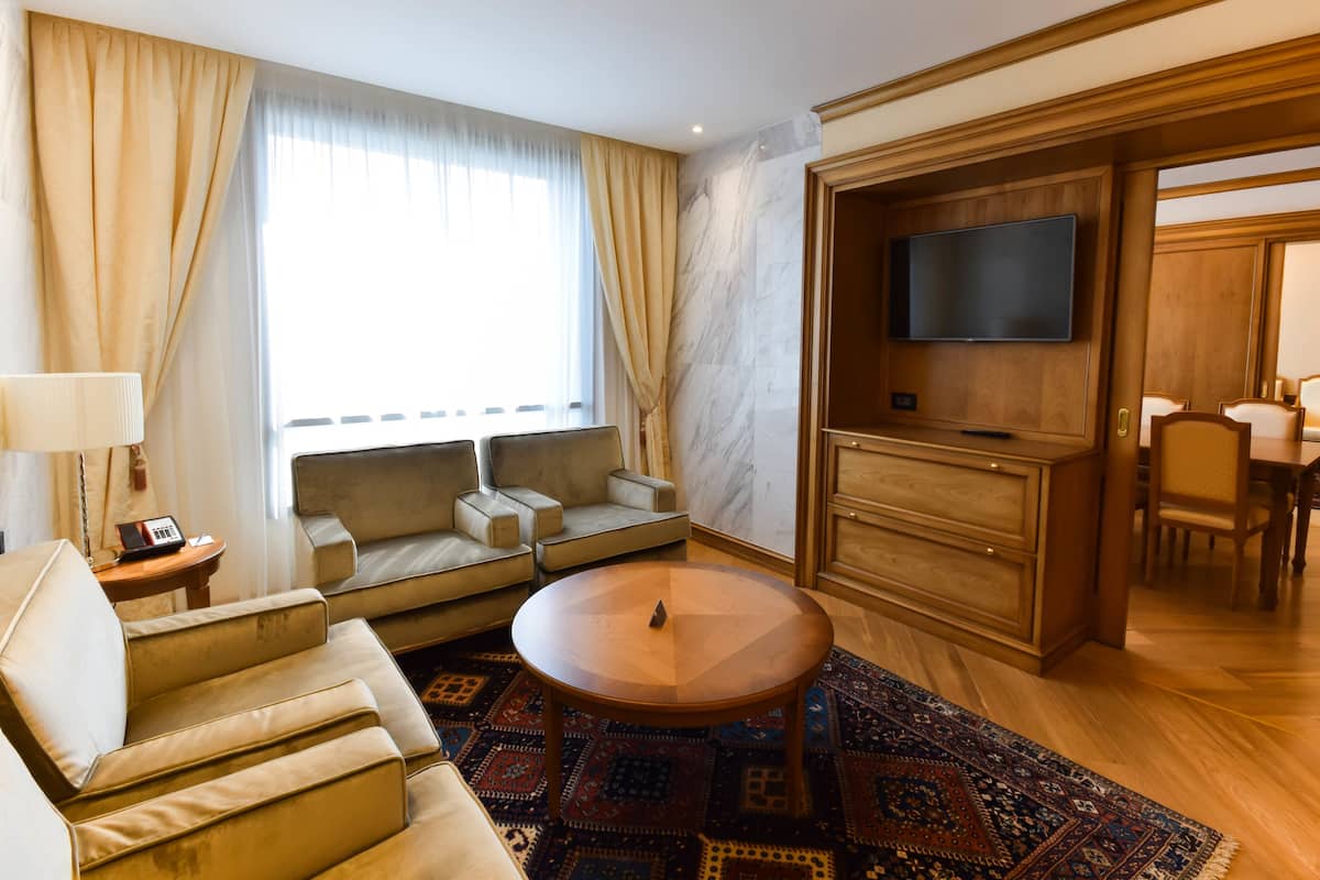 A modern hotel room with a sofa, armchair, round table, and a TV mounted on a wooden cabinet.