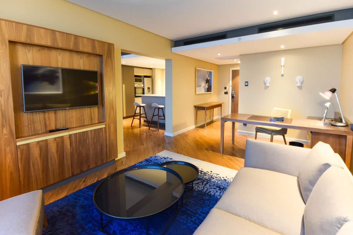 A modern living room with a flat-screen TV, a sofa, a coffee table, and a dining area with a kitchen in the background.
