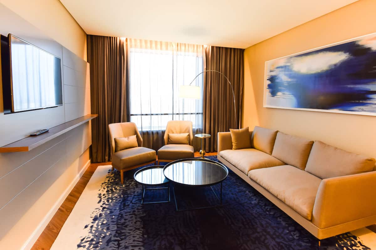 A modern hotel room with a sofa, two armchairs, a coffee table, a flat-screen TV, and a painting on the wall.