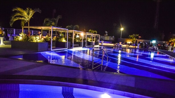 Un área de piscina iluminada con luces nocturnas, una zona de estar cubierta y palmeras.