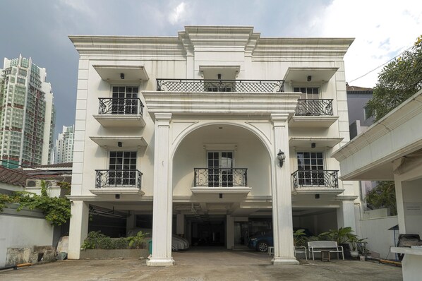A multi-story building with a central arched entrance, balconies with black railings, and a parking area in front.