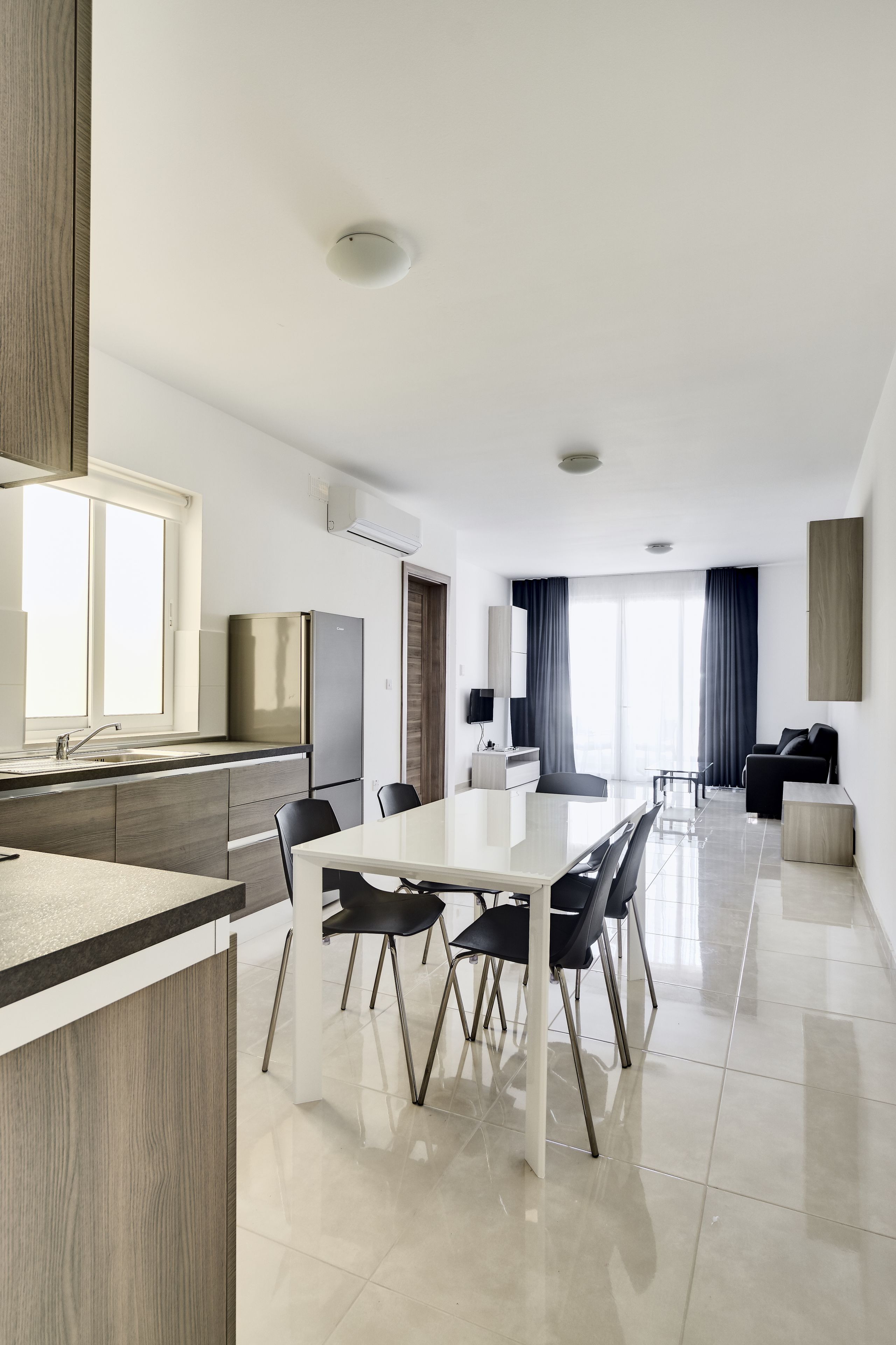 A modern kitchen with a dining area, a living room with a sofa and TV, and large windows with curtains.