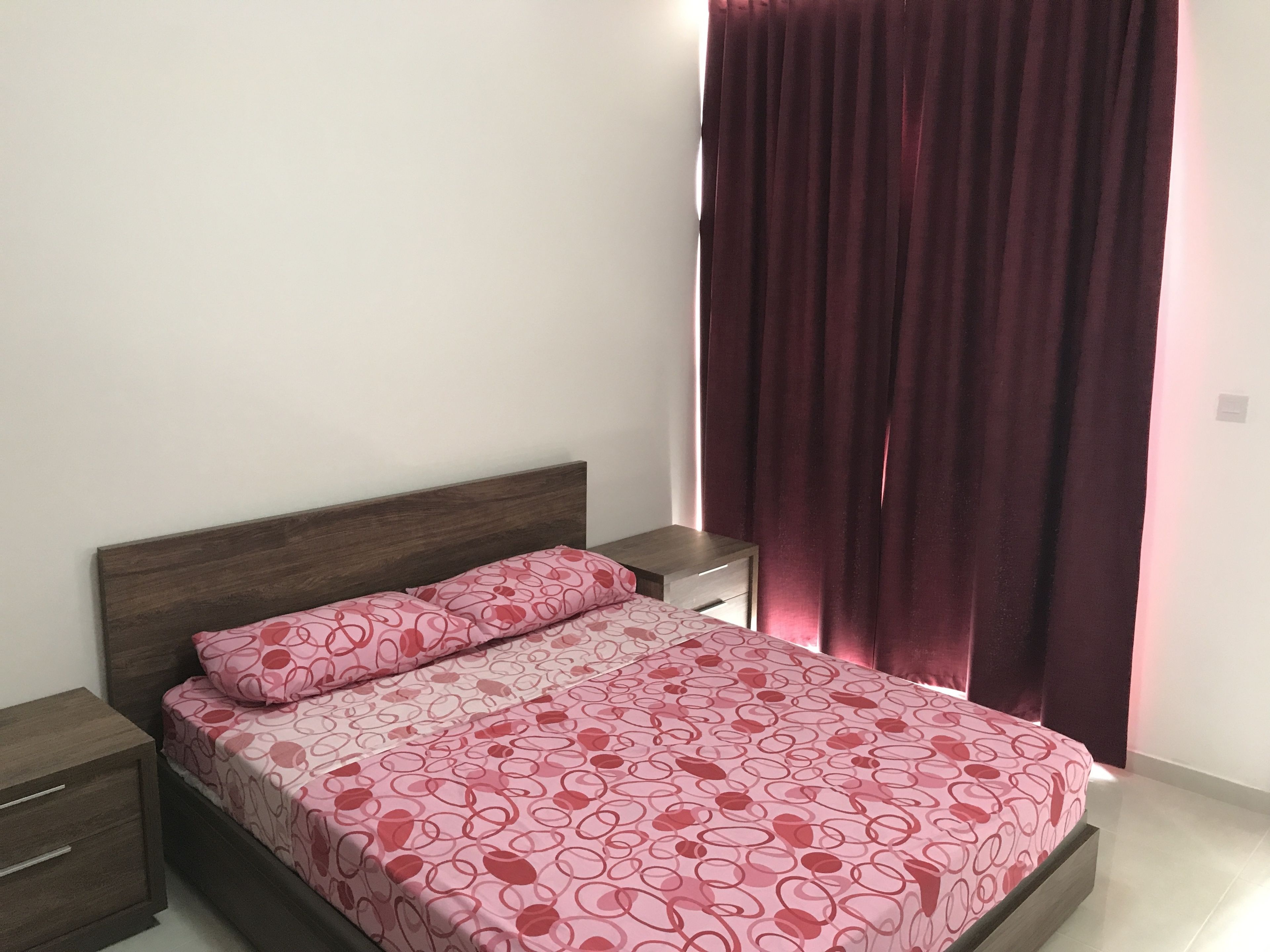 A bed with a patterned bedspread, a wooden headboard, a nightstand, and a dark curtain.