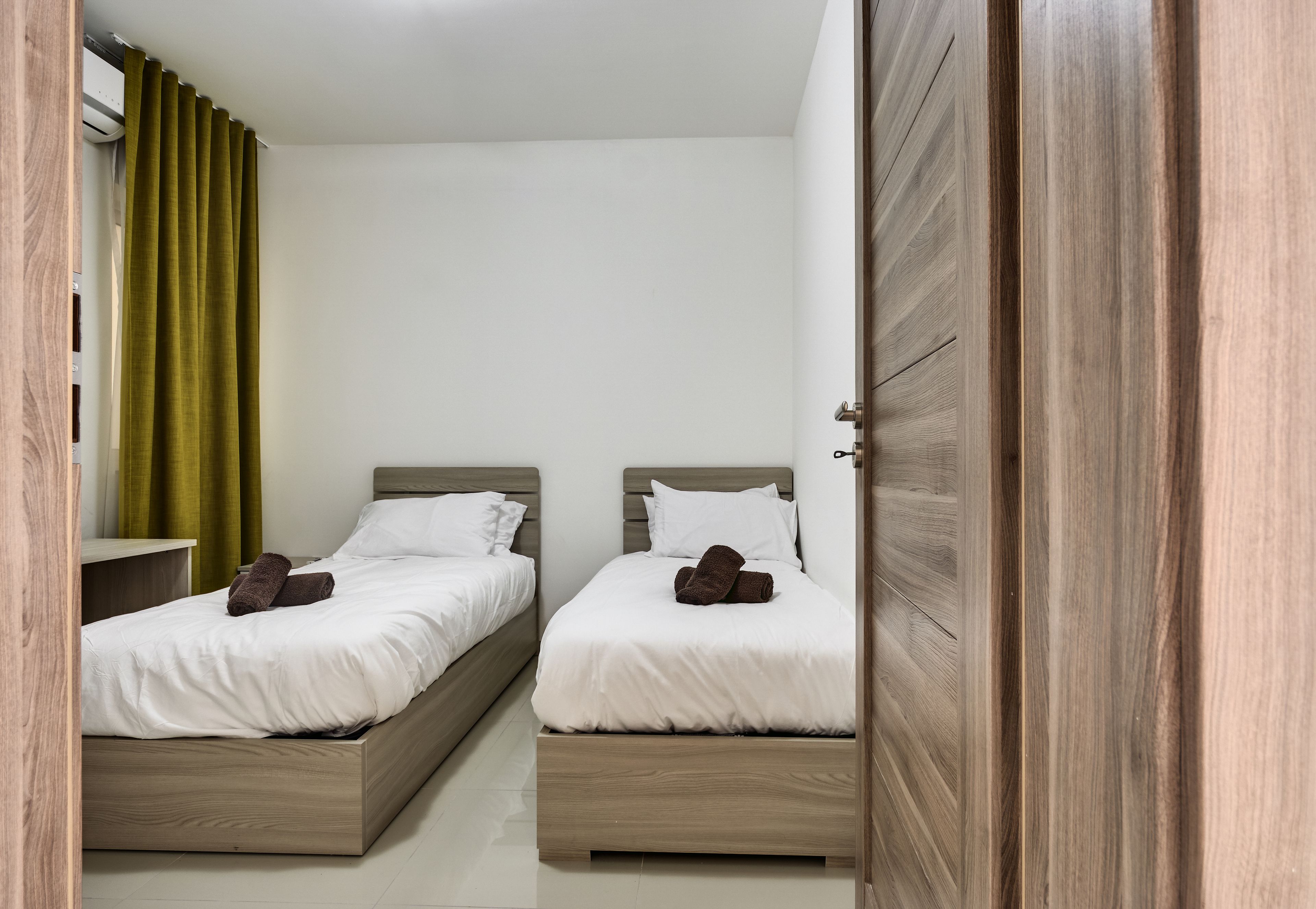 Ein Schlafzimmer mit zwei Betten, Holz-Kopfteilen, weißer Bettwäsche und einem grünen Vorhang.