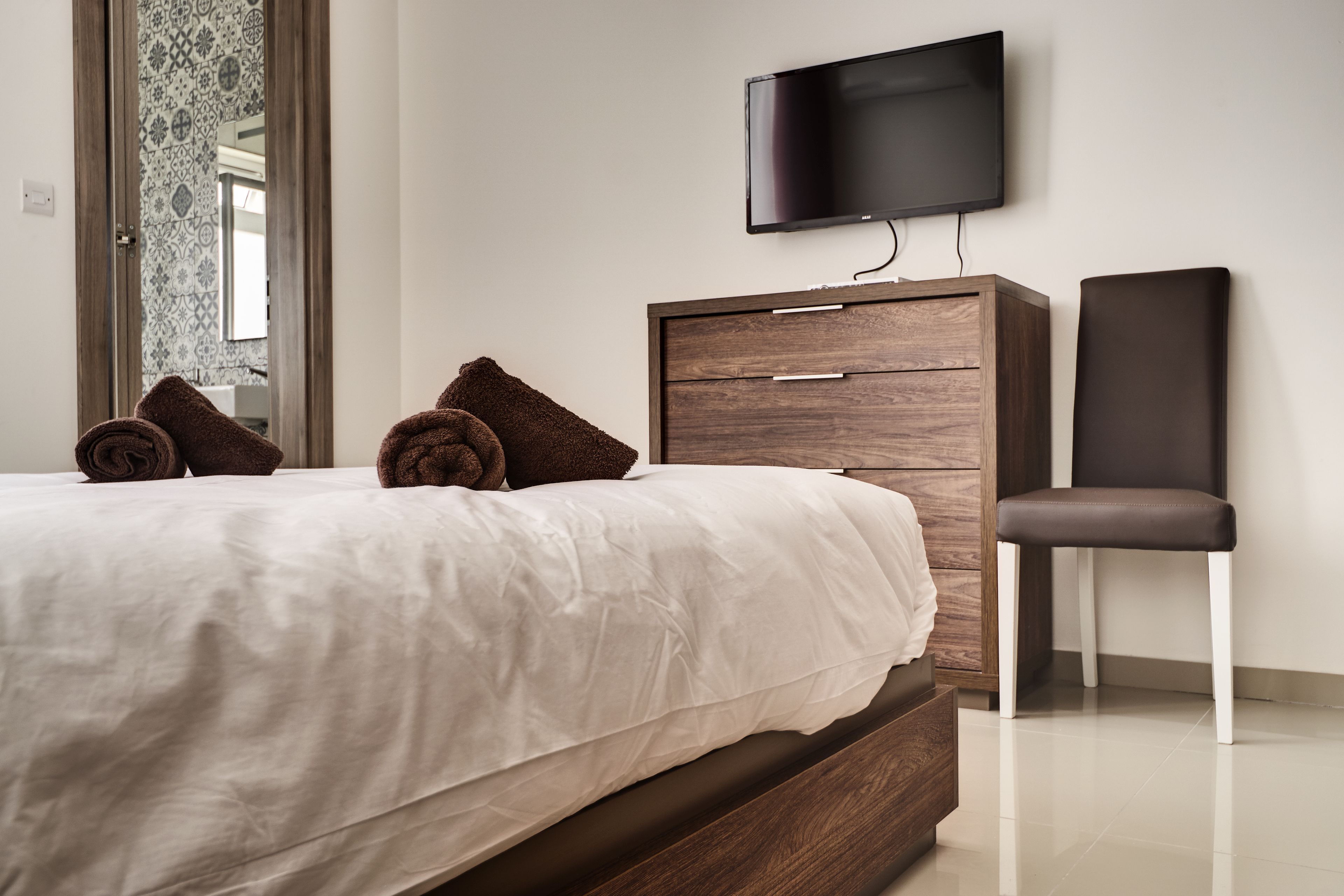 A bedroom with a bed, a TV mounted on the wall, a wooden dresser, and a chair.
