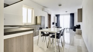 A modern kitchen with a central island, a dining area with a white table and black chairs, and a living area with a sofa and a TV. - The Village Apartments (St. Paul's Bay)