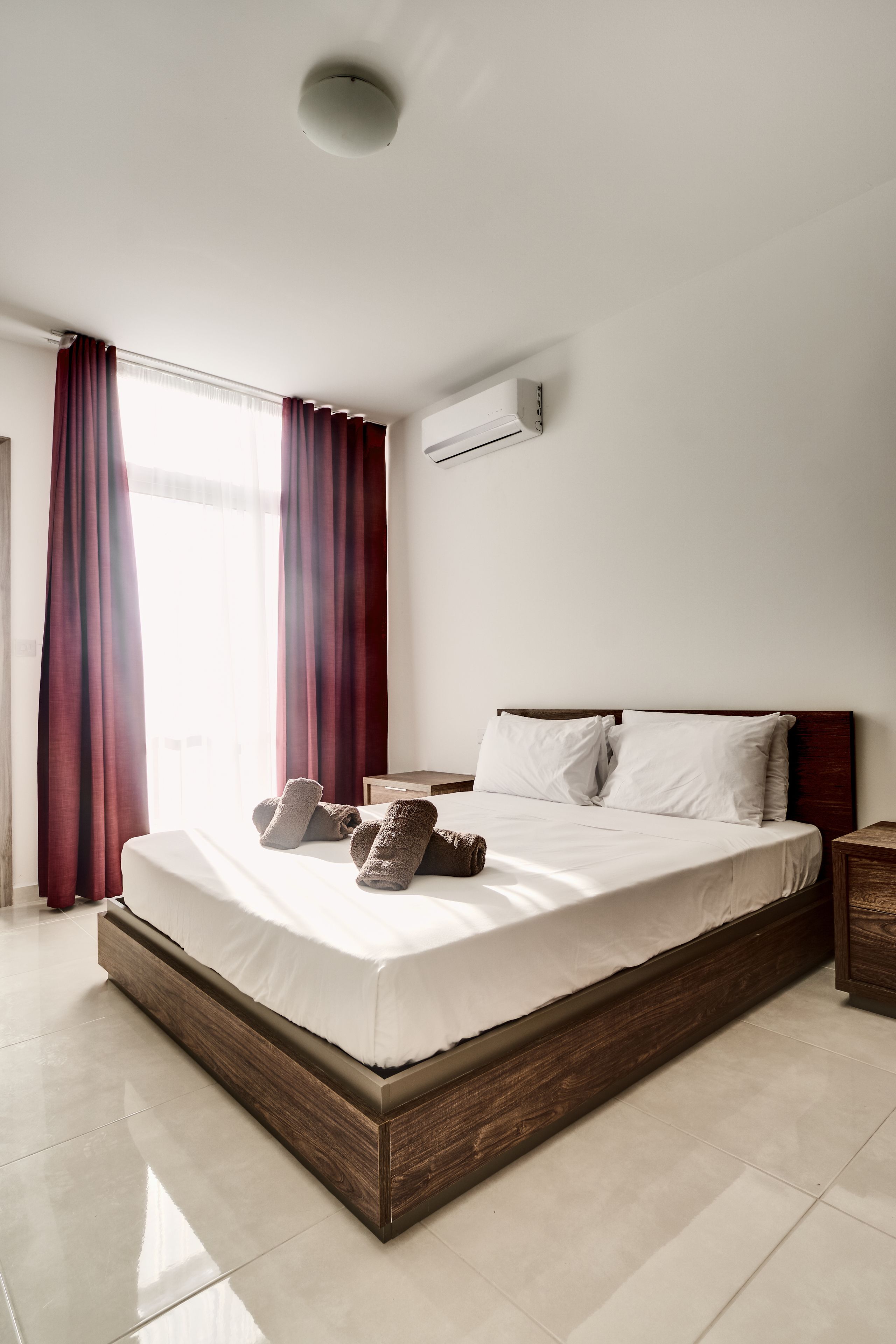 Una habitación de hotel moderna con una cama grande, cabecero de madera y un ventanal con cortinas rojas.