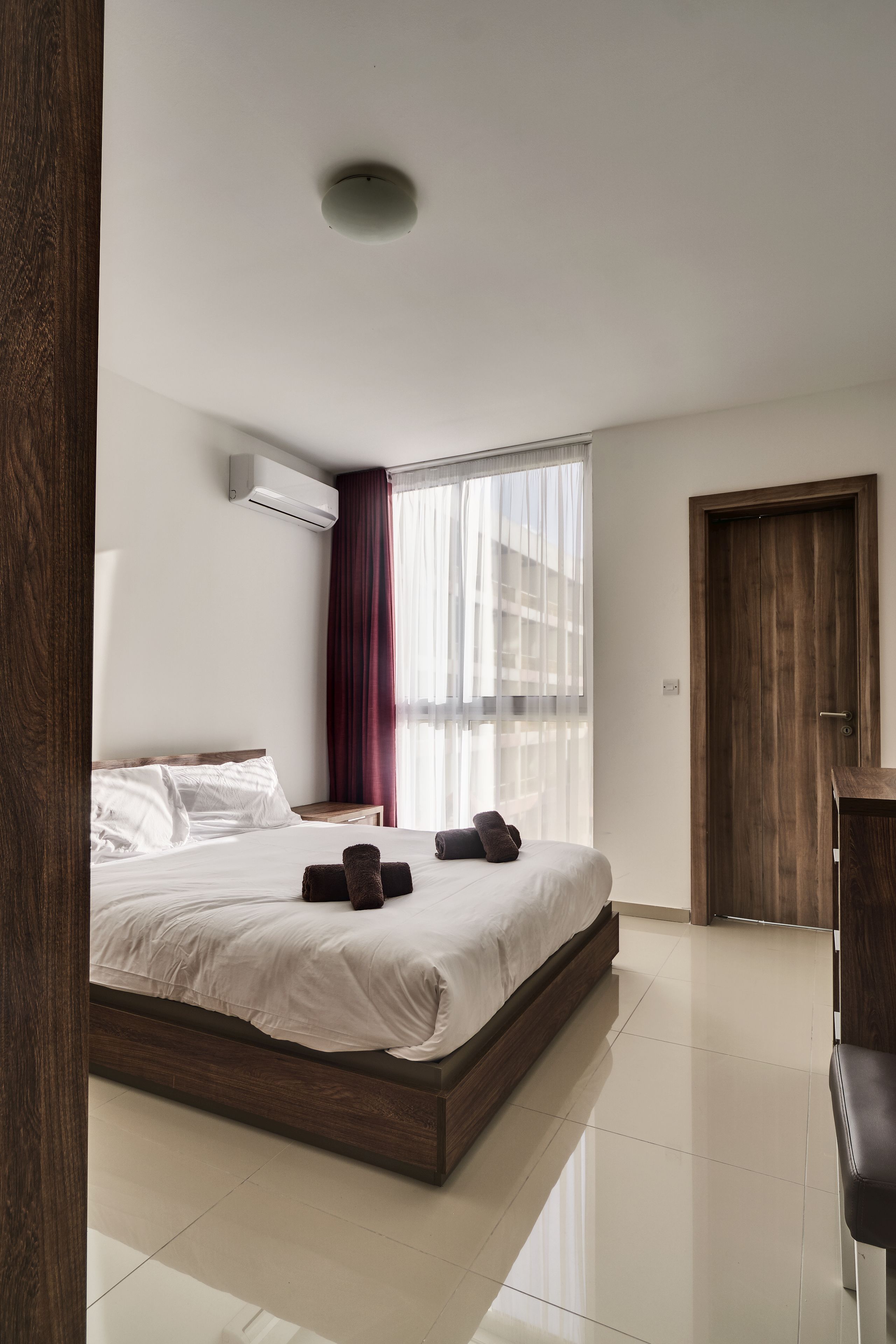 A modern hotel room with a large bed, a wooden door, and a window with sheer curtains.