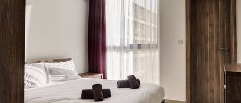 A modern hotel room with a large bed, a wooden door, and a window with sheer curtains.