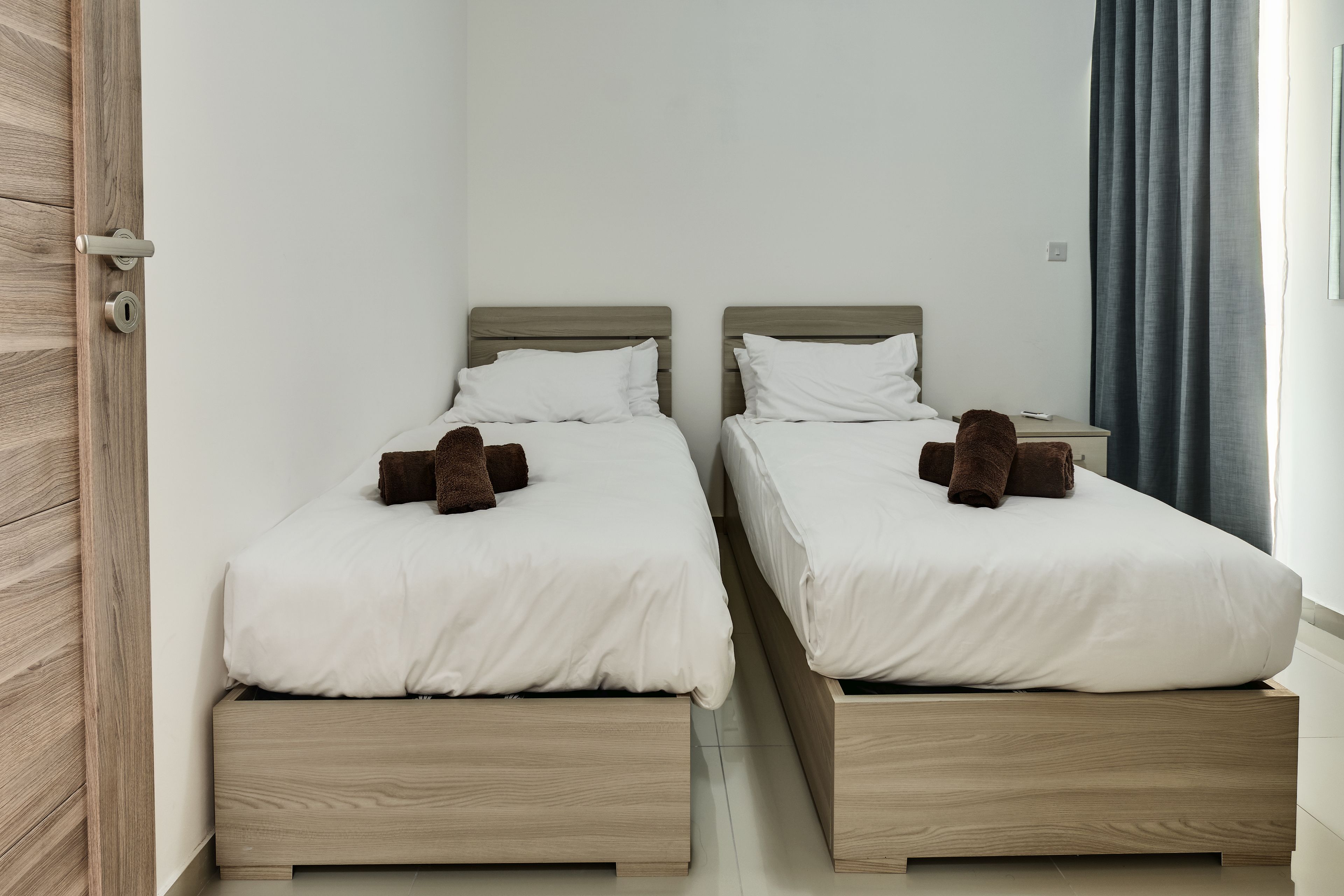 Une chambre avec deux lits, chacun doté d’une tête de lit en bois et de draps blancs. Il y a une porte en bois sur la gauche et une table de chevet avec une lampe sur la droite.