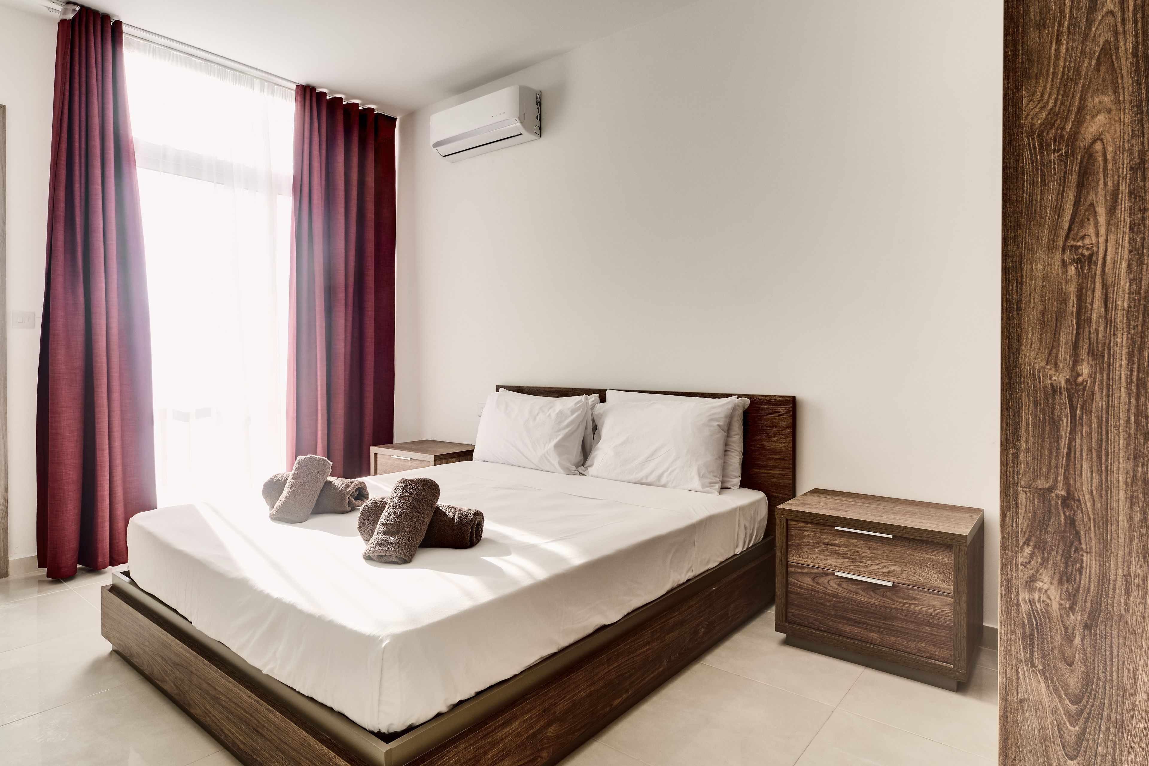 Moderni hotellihuone, jossa on puinen sängynrunko, valkoiset vuodevaatteet, yöpöytä ja puinen vaatekaappi.