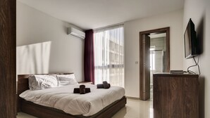A modern hotel room with a large bed, a wooden headboard, a TV mounted on the wall, and a large window with sheer curtains. - The Village Apartments (St. Paul's Bay)